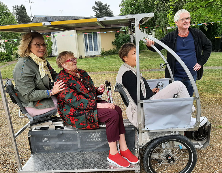 une personne âgée est assise sur le Vhelio, un vélo à deux places accompagnée d'une conductrice et d'une autre femme assise à l'arrière