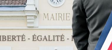 Vue de dos d'un élu portant une écharpe tricolore (bleu, blanc, rouge) devant le fronton d'une mairie. Sur la façade du bâtiment, on peut lire les mots « MAIRIE » et la devise républicaine « LIBERTÉ - ÉGALITÉ ». L'image illustre la proximité et l'engagement des élus locaux.