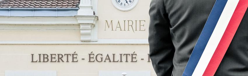 Vue de dos d'un élu portant une écharpe tricolore (bleu, blanc, rouge) devant le fronton d'une mairie. Sur la façade du bâtiment, on peut lire les mots « MAIRIE » et la devise républicaine « LIBERTÉ - ÉGALITÉ ». L'image illustre la proximité et l'engagement des élus locaux.