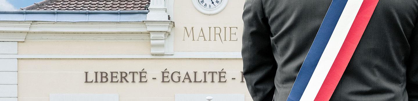 Vue de dos d'un élu portant une écharpe tricolore (bleu, blanc, rouge) devant le fronton d'une mairie. Sur la façade du bâtiment, on peut lire les mots « MAIRIE » et la devise républicaine « LIBERTÉ - ÉGALITÉ ». L'image illustre la proximité et l'engagement des élus locaux.