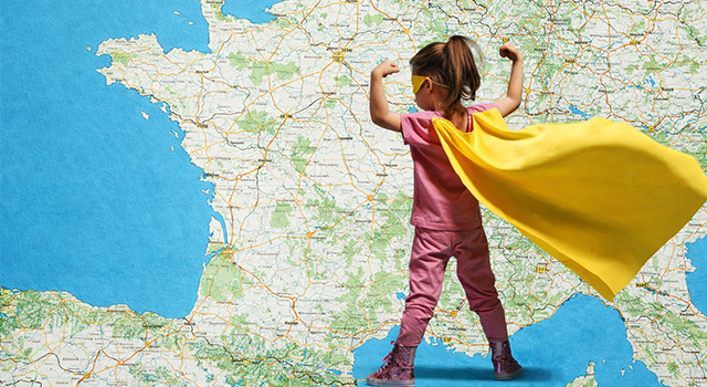 Illustration montrant une petite fille de dos, déguisée en super-héroïne avec une cape jaune et un loup. Elle se tient debout, les bras levés montrant ses muscles, devant une immense carte de France détaillée. L'image symbolise la force et l'avenir des territoires.
